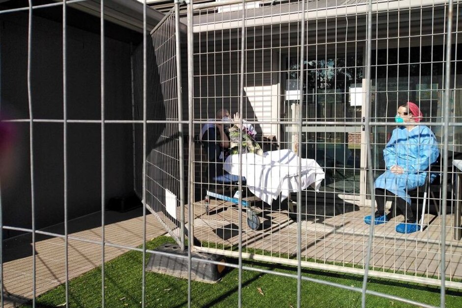 Elderly person in bed and nurse in scrubs sitting behind fence