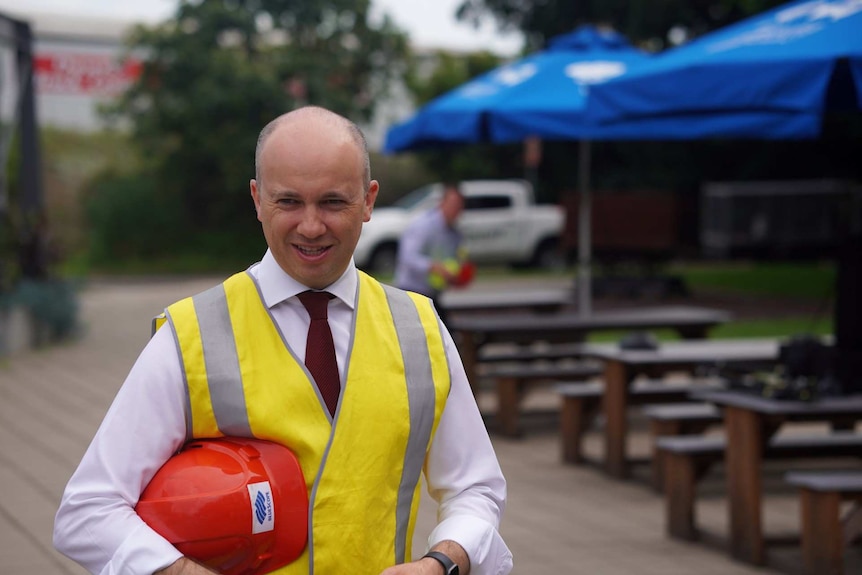 A bald man wearing a hi-vis vest over a shirt and tie holds a hard hat in the crook of his arm.