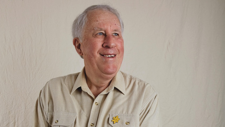 A man smiles and looks off camera. He is an older man in a beige shirt.