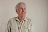 A man smiles and looks off camera. He is an older man in a beige shirt.