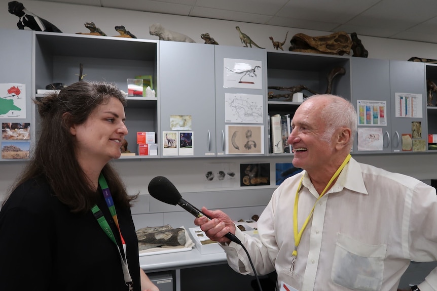 Man with a microphone interviews a woman in a room with dinosaur bones on a shelf in the background.