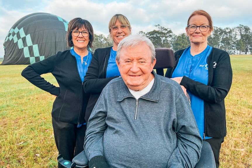 A man in a wheelchair smiles at the camera, while three women (his carers) stand behind him, smiling.