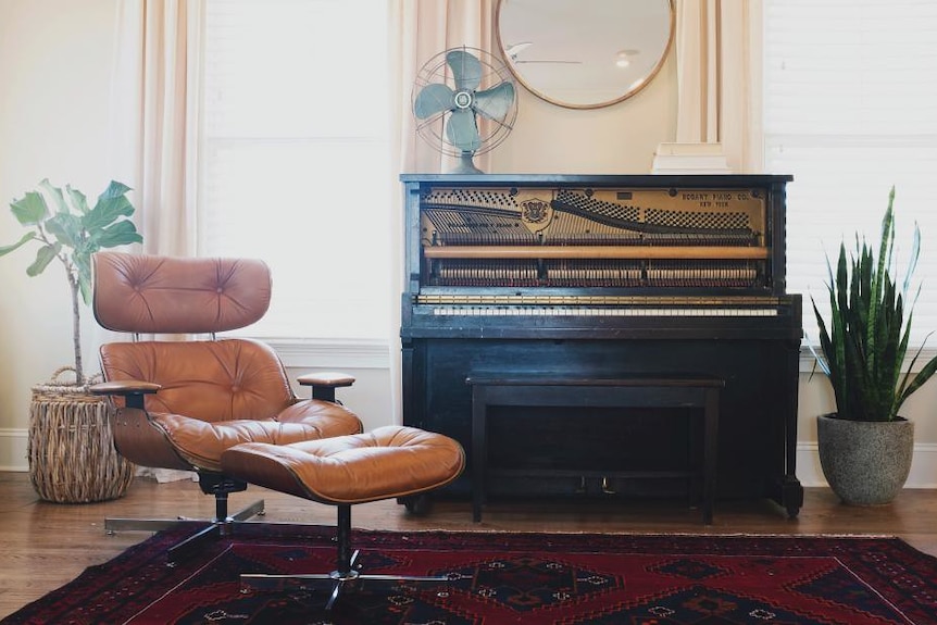 A styled living room scene with an upright piano showing the strings and hammers, a brown Eames lounge chair and some plants.