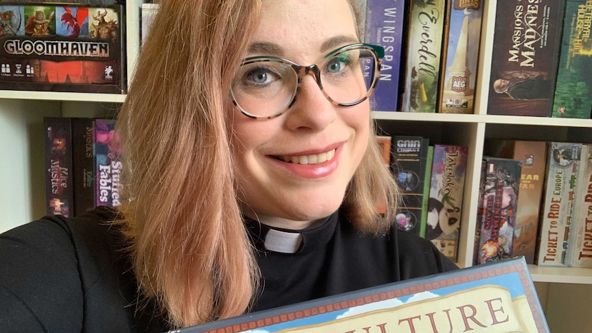 Uniting Church minister The Reverend Amanda Hay wearing a clerical collar holding her favourite board game, Viticulture.