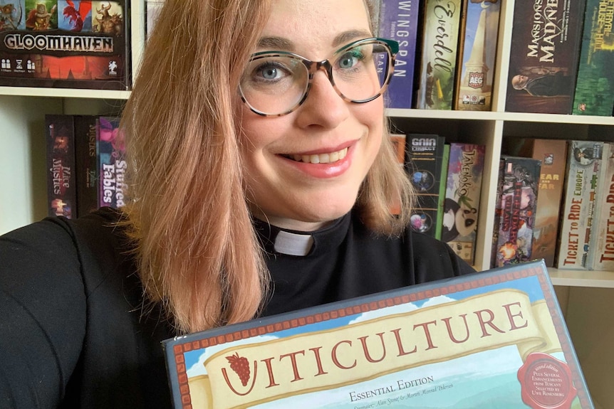 Uniting Church minister The Reverend Amanda Hay wearing a clerical collar holding her favourite board game, Viticulture.