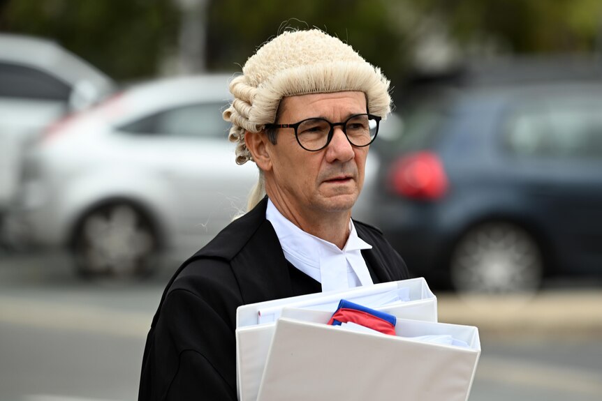 Mr Drumgold walks along, wearing a wig and carrying a stack of papers and documents.