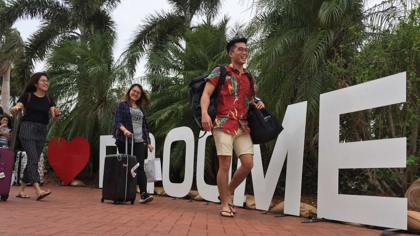 Three people with suitcases walk past a Broome sign smiling