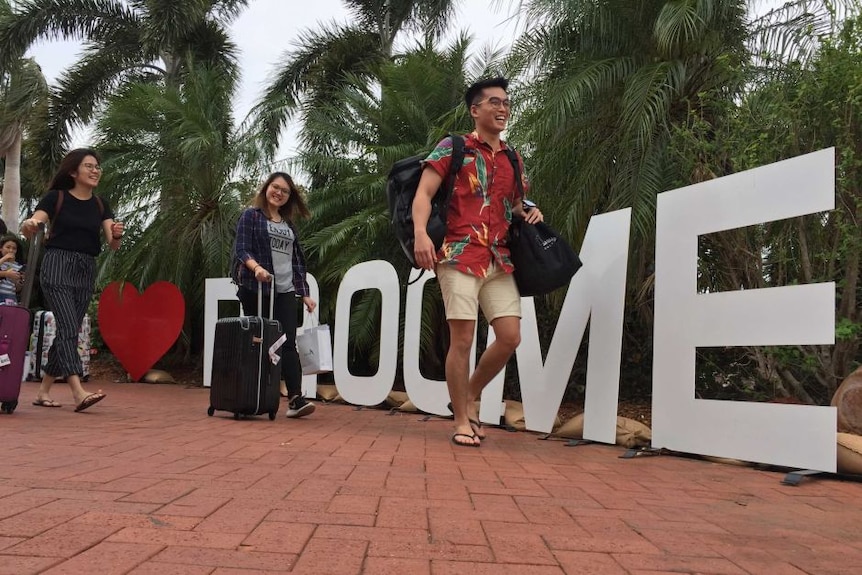 Three people with suitcases walk past a Broome sign smiling