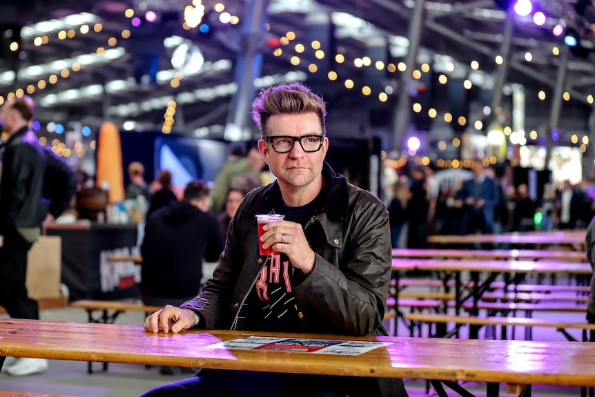 Man wearing black rimmed glasses and black jacket holds beer sitting at wooden table amid dimly lit beer hall