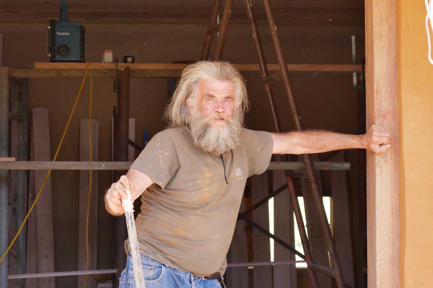 A man with long white hair and a beard stands in the doorway of a building under construction.