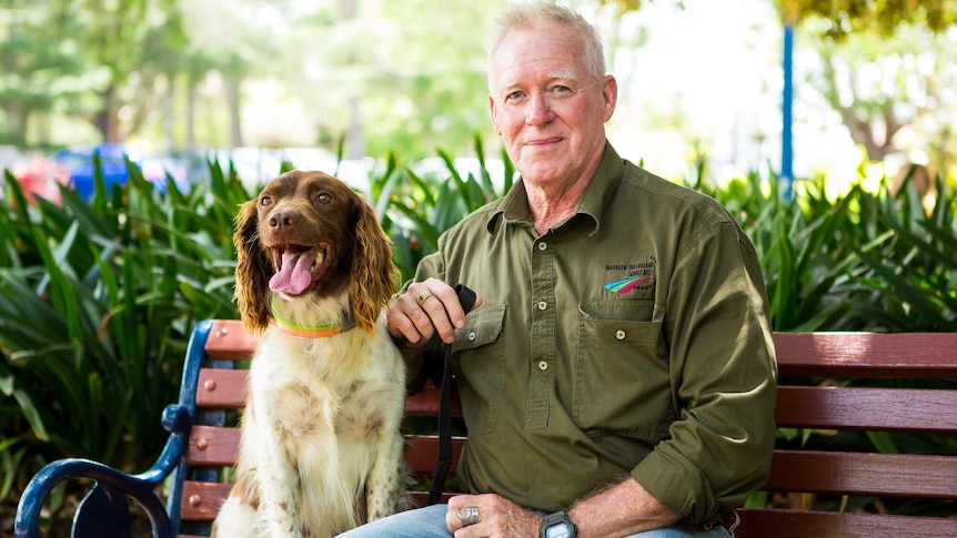 A smiling middle-aged man in an olive shirt and jeans and a medium-sized brown and white dog sit on a park bench.