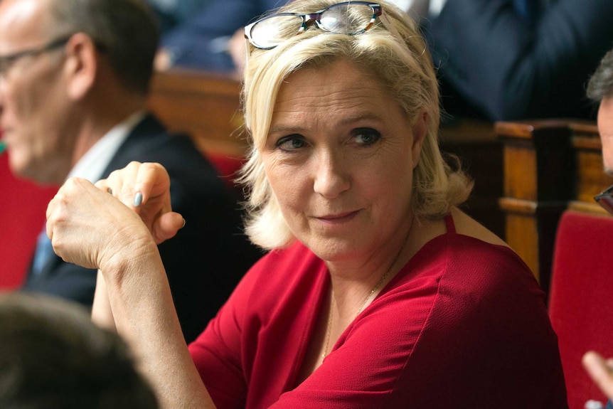 Marine Le Pen looks off the left while wearing a red dress and her reading glasses on her head.