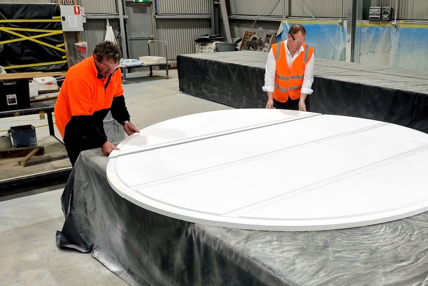 Two men in high vis place large white piece of fibreglass on table to form circular floor of igloo.