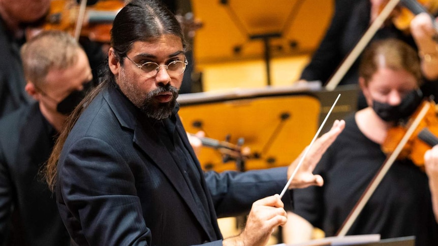 Aaron, wearing all black, conducts the Melbourne Symphony Orchestra, turning his head to the right to look at the players