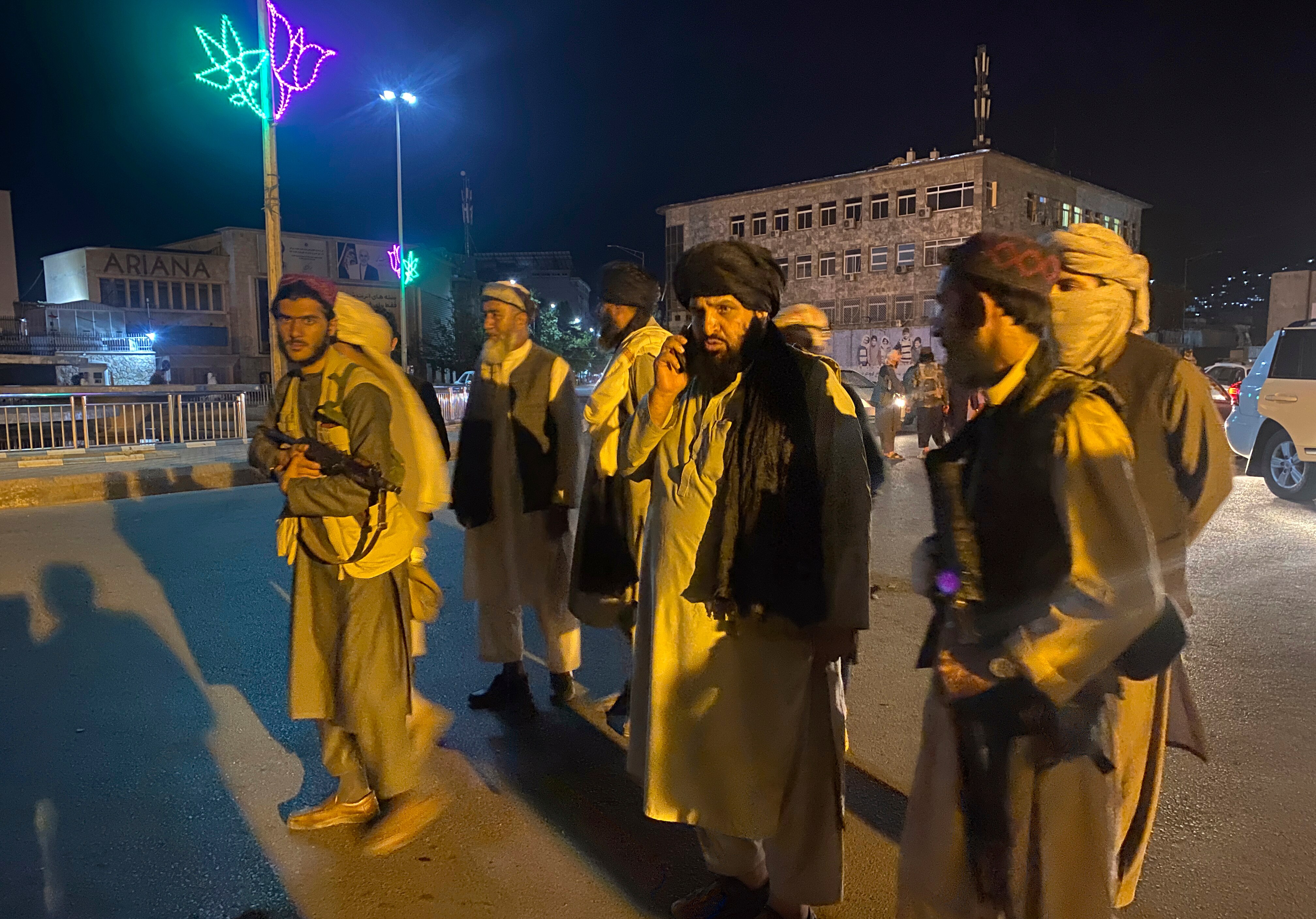Seven men, some armed, wearing traditional Afghan clothing walk along a street at night.