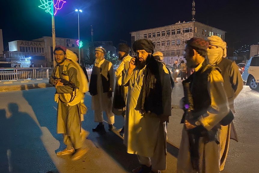 Seven men, some armed, wearing traditional Afghan clothing walk along a street at night.