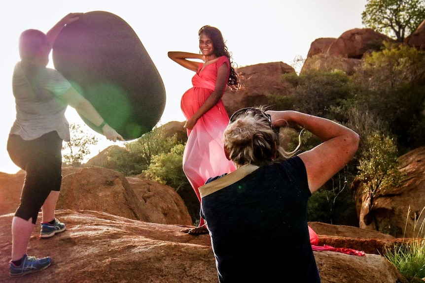 A pregnant woman wearing a pink dress stands on boulders posing for a photographer while another woman holds a reflector
