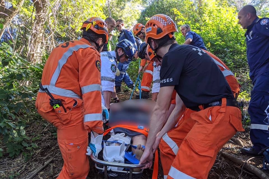 A group of people in high-vis SES, police rescue and NSW ambulance uniforms carry a woman (face blurred) on stretcher in bush.