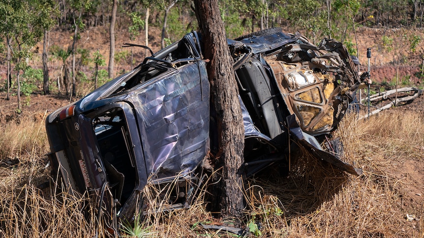 A blue landcruiser lies on its side against a tree. It is extensively damaged. 