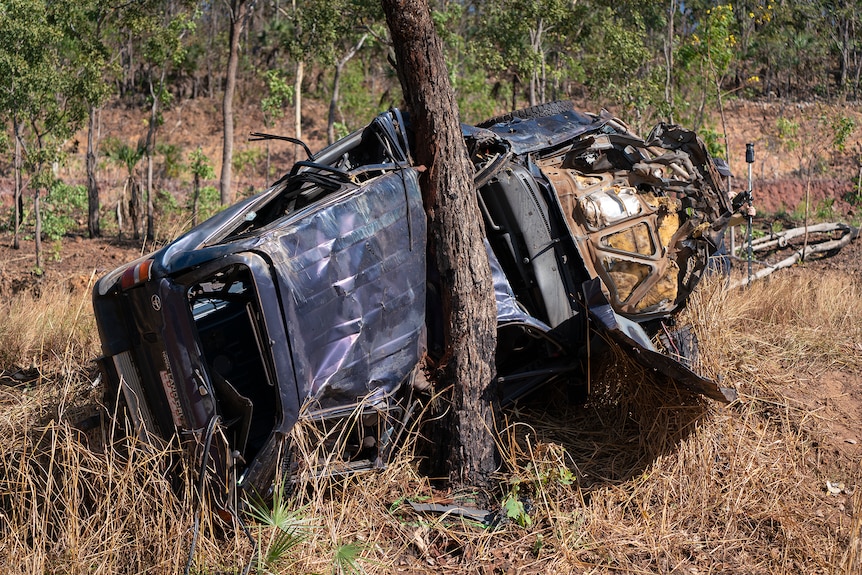 A blue landcruiser lies on its side against a tree. It is extensively damaged. 