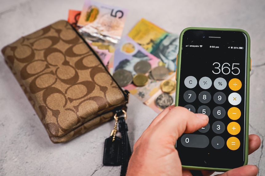 A man types numbers 365 into a phone calculator with cash and a wallet in the background