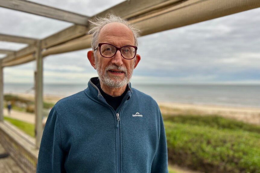 A man with grey hair and glasses looks directly at the camera, with a beach in the background