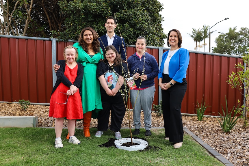 A group of brightly dressed people stand smiling behind a newly planted tree.