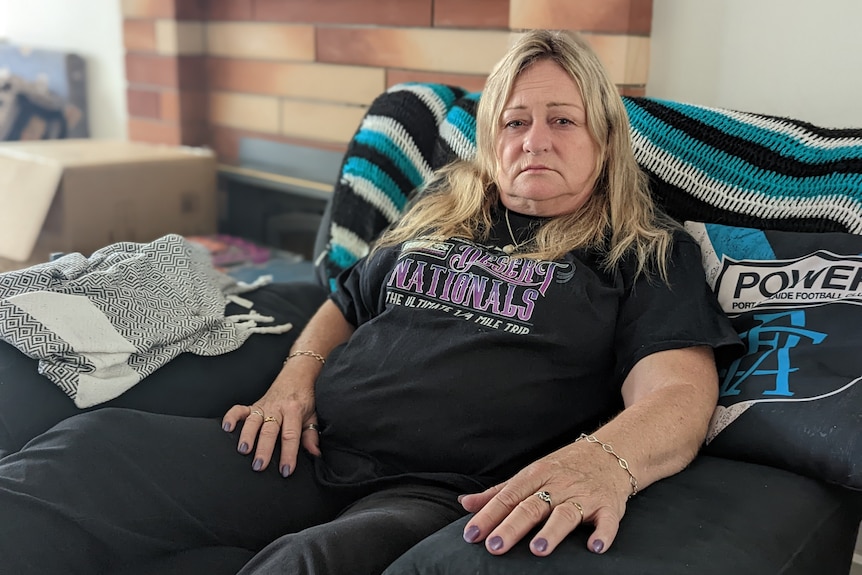 A woman with long blonde hair, wearing a black T-shirt, sits on a couch