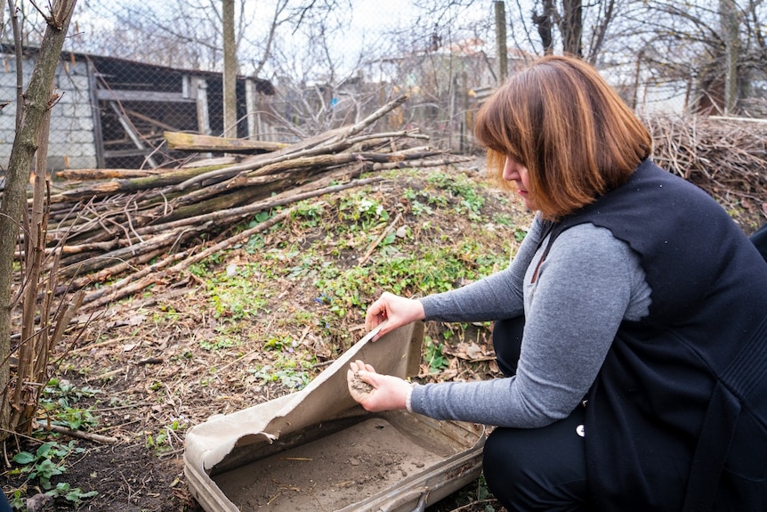 A woman kneeling on the ground outside, leaning over an old suitcase, and holding soil and debris in her hand