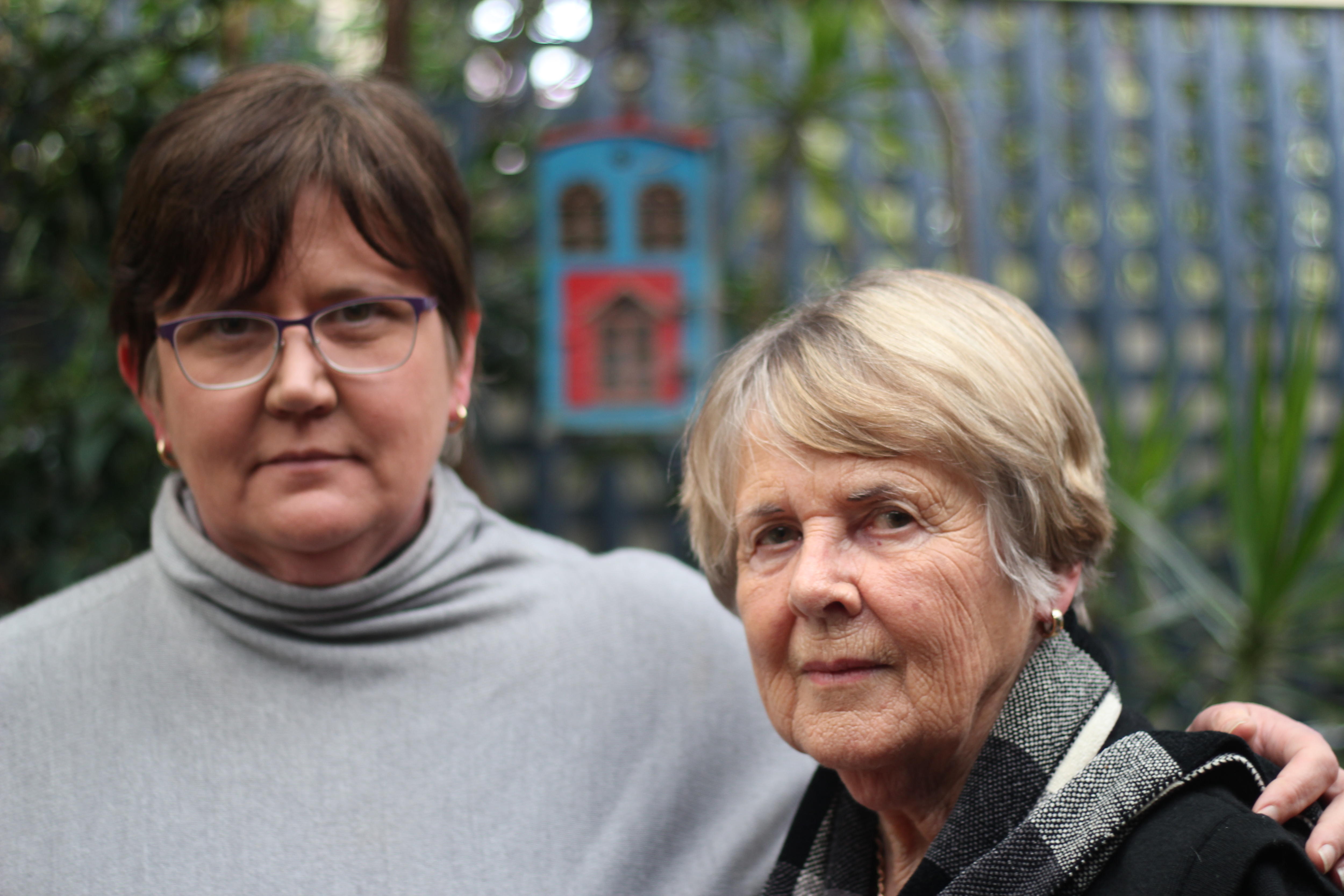 Two women stand in a home garden looking at a camera