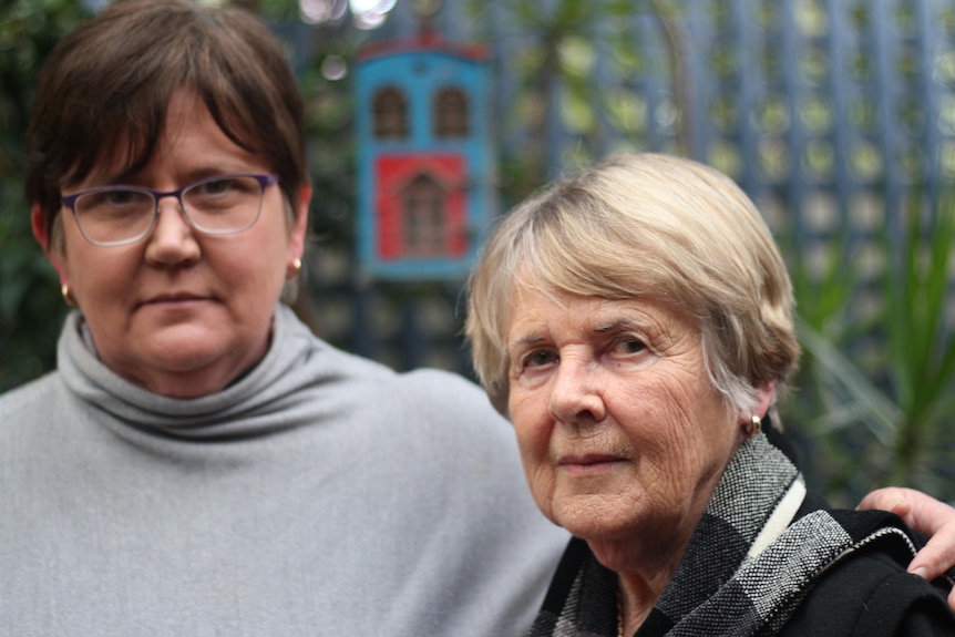 Two women stand in a home garden looking at a camera