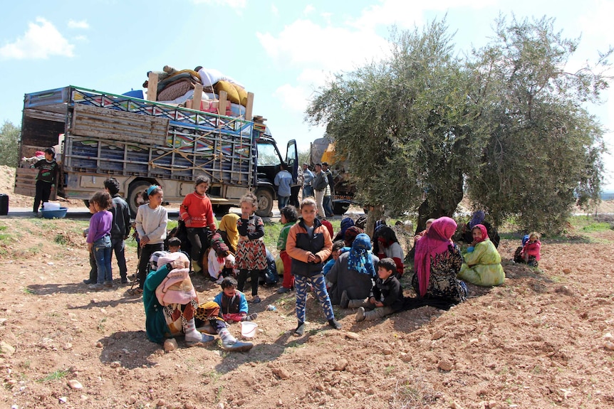 Women and children resting in a field, in front of a truck loaded with luggage