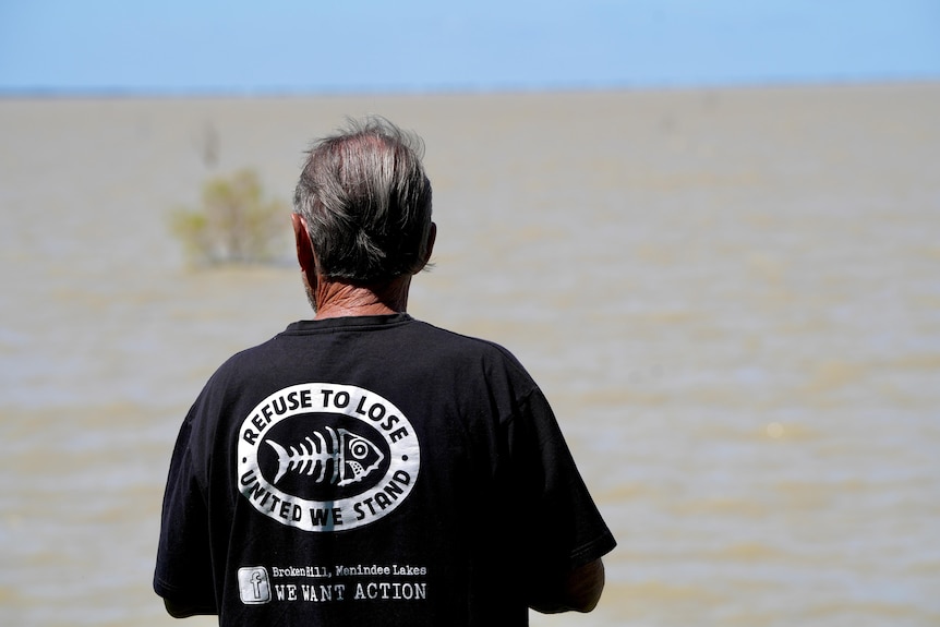 A man in a blakc tshirt which reads 'refuse to lose' looking out at a full lake Menindee