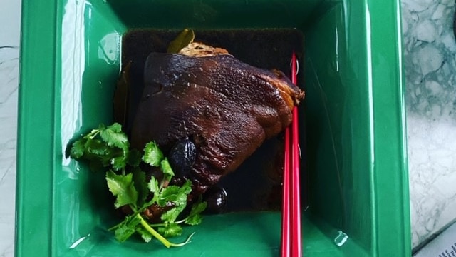 A deep caramel browned pork hock sitting in a dark sauce with coriander garnish and red chopsticks in a green bowl.