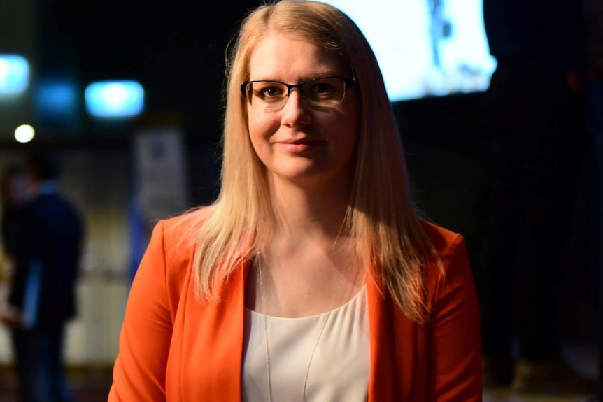 A close-up of a blonde woman wearing an orange jacket.