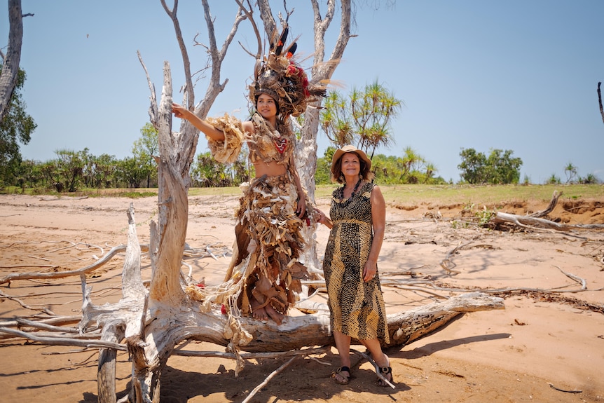 Two women stand on and next to a tree branch on a beach. One is wearing an elaborate costume