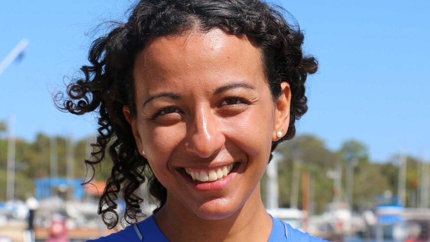 A headshot of a woman in a blue top smiling with boats in the background.