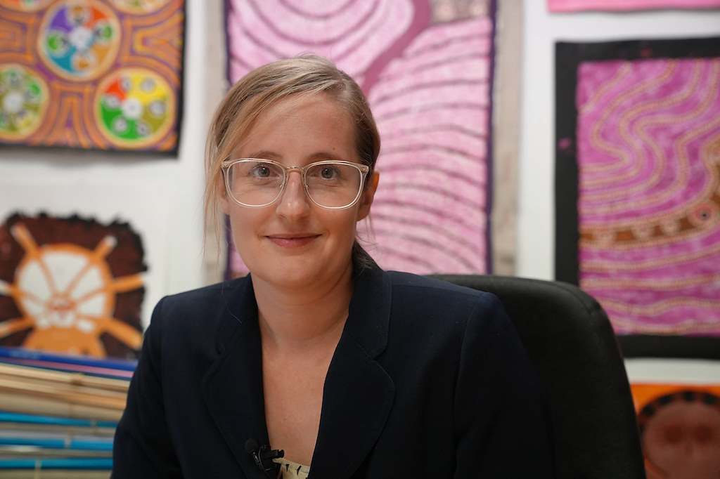 Sally wears glasses and smiles as she sits in front of Aboriginal artwork