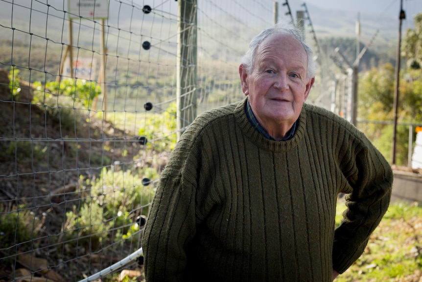 A men wearing a green knitted jersey leans on a walking stick in front of a high electric fence.