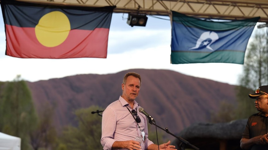 Nigel Scullion stands in front of Uluru under Aboriginal and Torres Strait Islander flags.