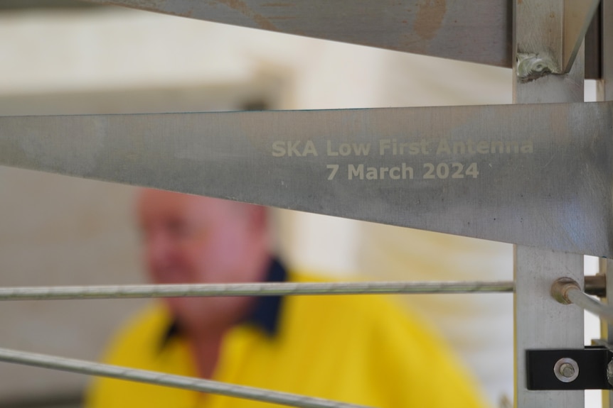 A man in high-vis wear standing behind a plaque that reads 'SKA Low first antenna March 7 2024'