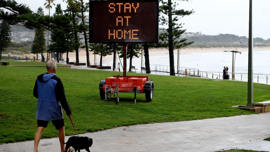 A man walking a dog near the beach walks part an electronic sign that reads: "Stay at home"