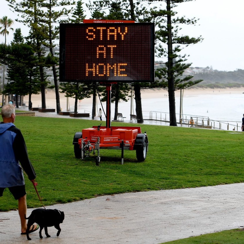 A man walking a dog near the beach walks part an electronic sign that reads: "Stay at home"