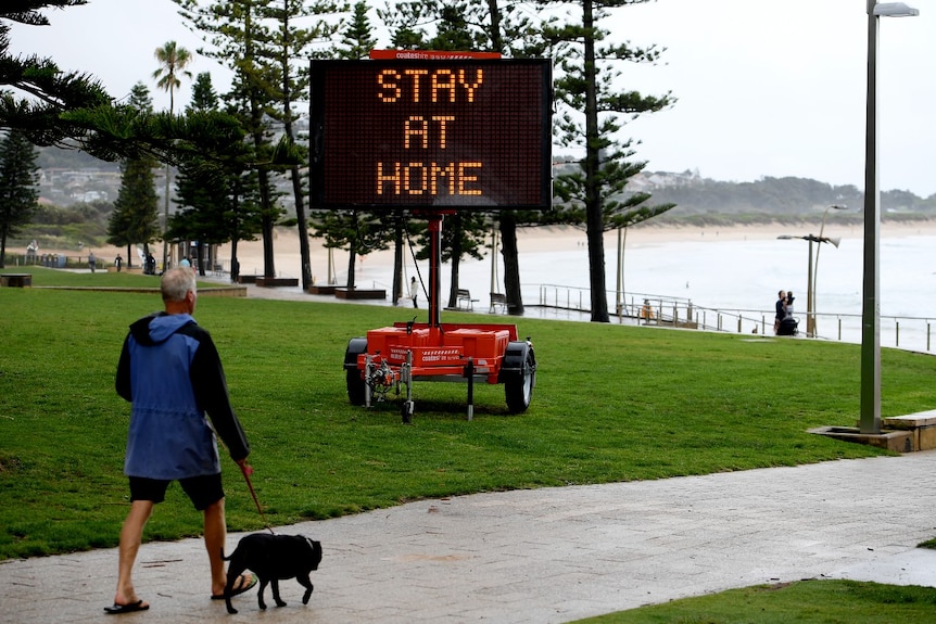 A man walking a dog near the beach walks part an electronic sign that reads: "Stay at home"