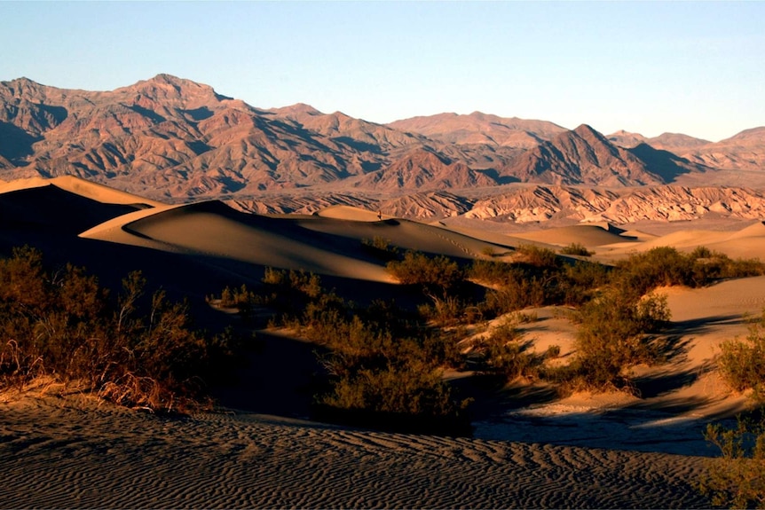 Sand dunes sit in front of a spectacular, rocky mountain range.