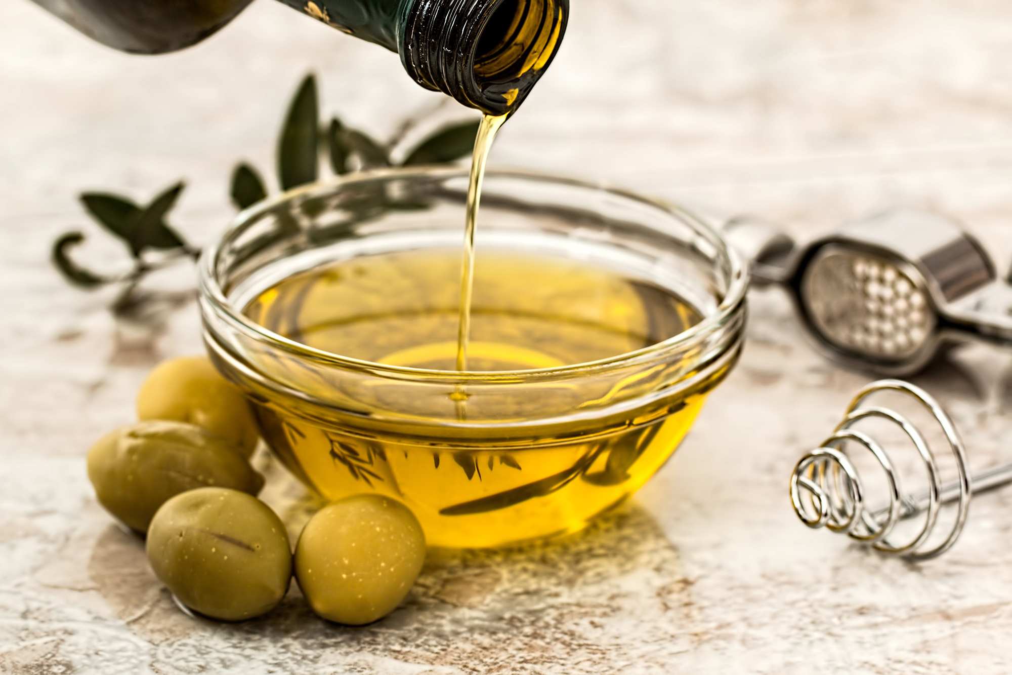 Close-up of olive oil being poured into a small glass dish, next to olives and olive branch.
