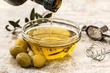 Close-up of olive oil being poured into a small glass dish, next to olives and olive branch.