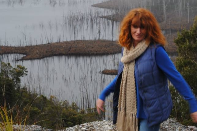 A woman with red hair wearing blue and a tan scarf smiles at the camera with lake in background