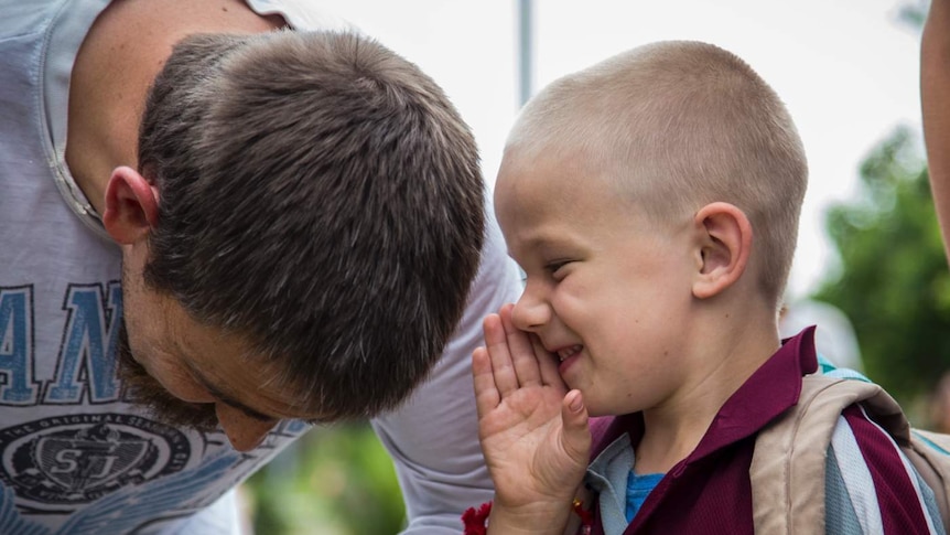 A young boy holds his hand to his mouth and speaks to an older man, who is bending down to listen