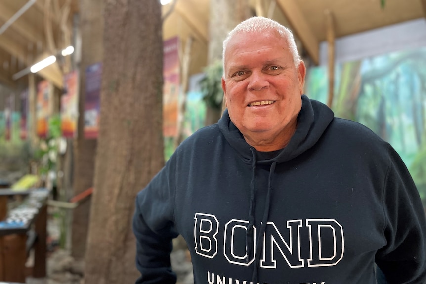 A man with white hair wearing a navy Bond University jumper standing in front of an indoor forest setting.